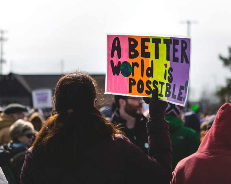A woman holding a colorful sign that says “A Better World is Possible” while in a crowd at a protest