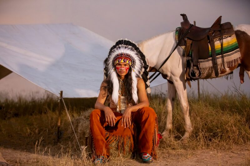 American indian man with headdress and makeup with his horse.