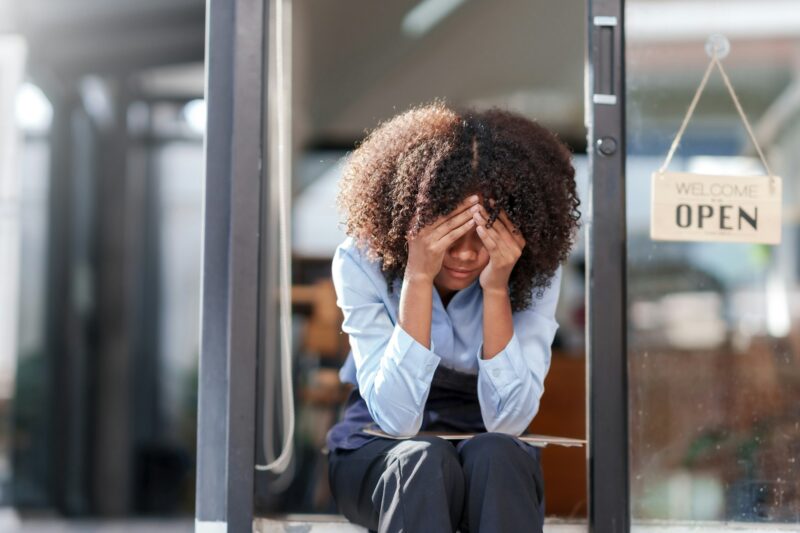 Female shopkeeper sitting stressed out at the store entrance frustrated by the economic impact.