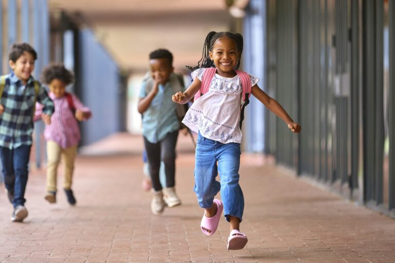 Group Of Multi-Cultural Elementary School Pupils Running Along Walkway Outdoors At School