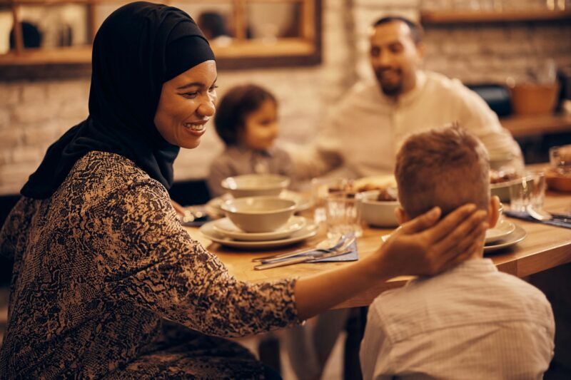 Happy Black Islamic woman enjoying with her family during meal at dining table.