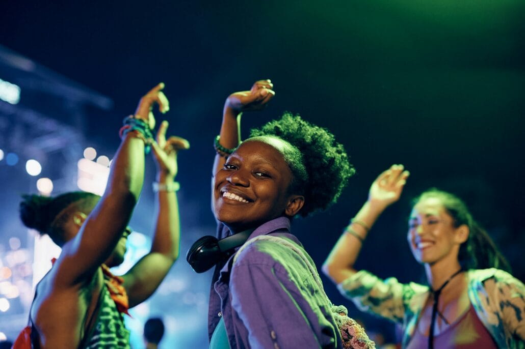 Happy black woman dancing while attending summer music festival at night and looking at camera.