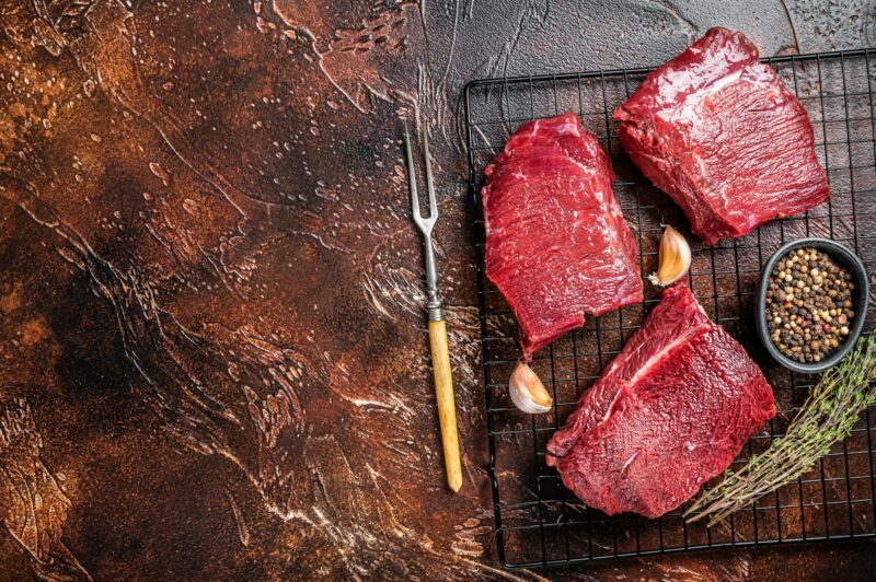 Raw Wild Game Meat of Venison dear ready for cooking. Dark background. Top view. Copy space
