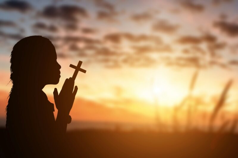 Silhouette of little girl holding christian cross in hands to praying for blessing from god at