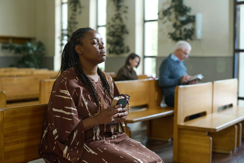 Young Christian black woman in casual dress sitting on bench and praying