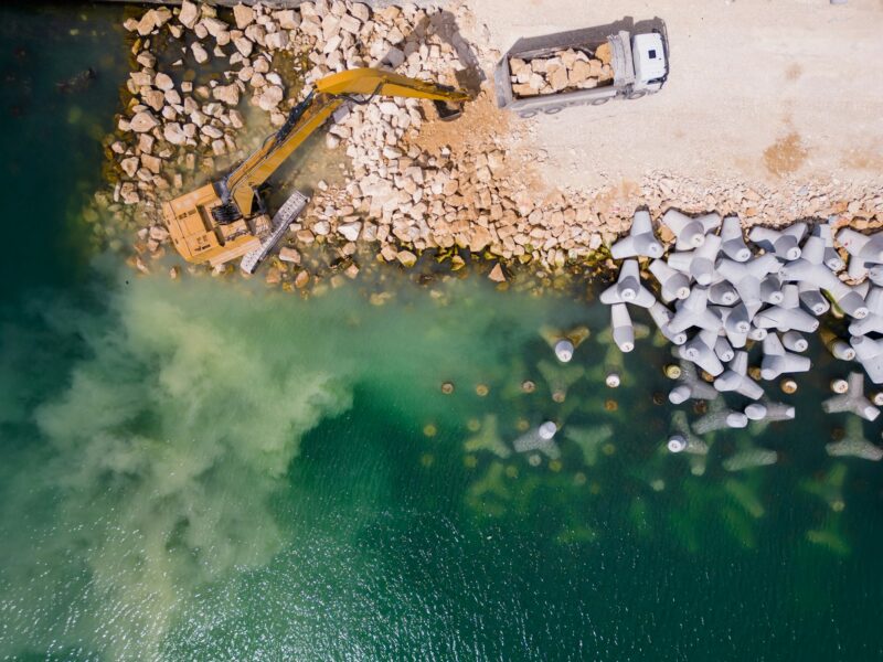 An excavator diligently constructs a dock or breakwater in the sea, its mighty arm reaching out from