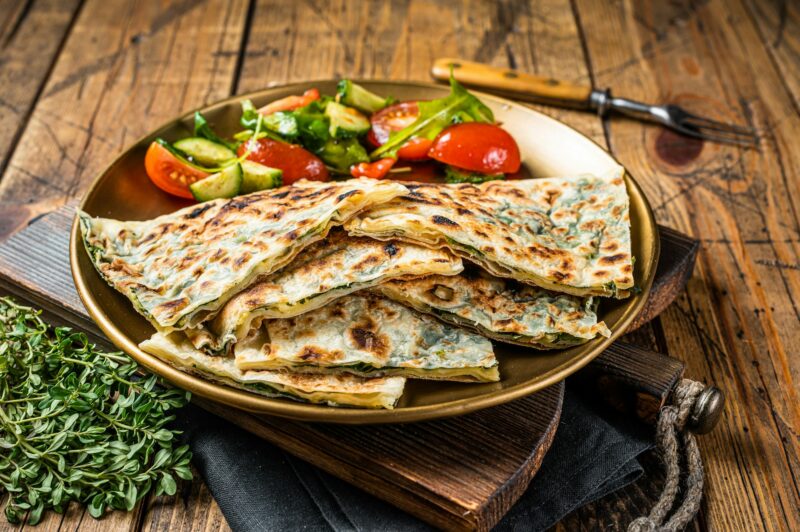 Gozleme flatbread with greens and vegetable salad on garnish. Wooden background. Top view