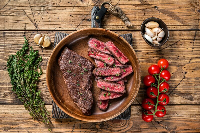 Grilled sliced top blade or denver beef meat steak in a wooden plate with herbs.