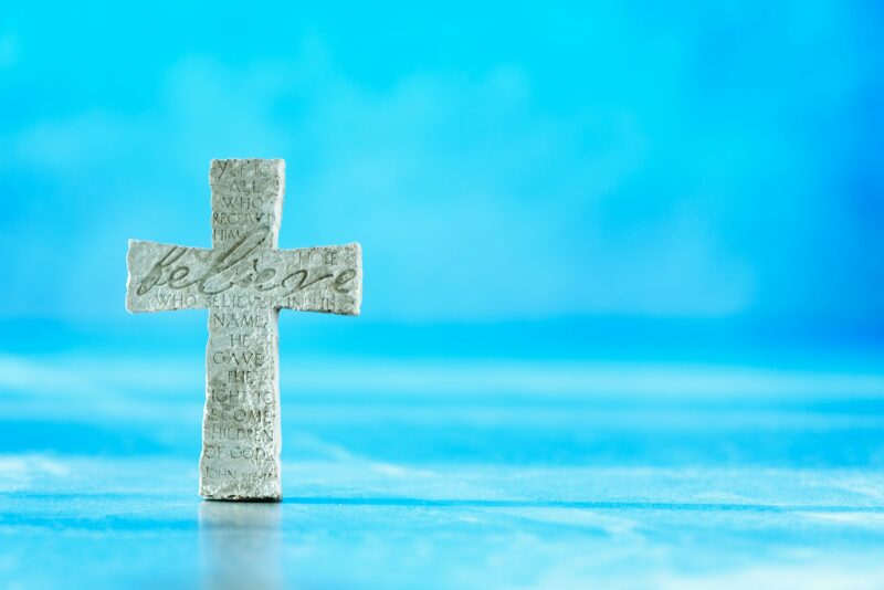 Stone cross with inscription Believe on blue background, Copy space. Christian backdrop. Biblical
